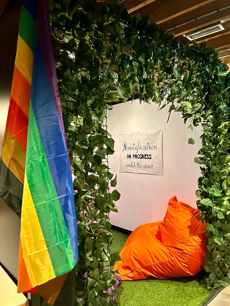 A pride flag and comfortable nook with sign stating 'beautification in progress'